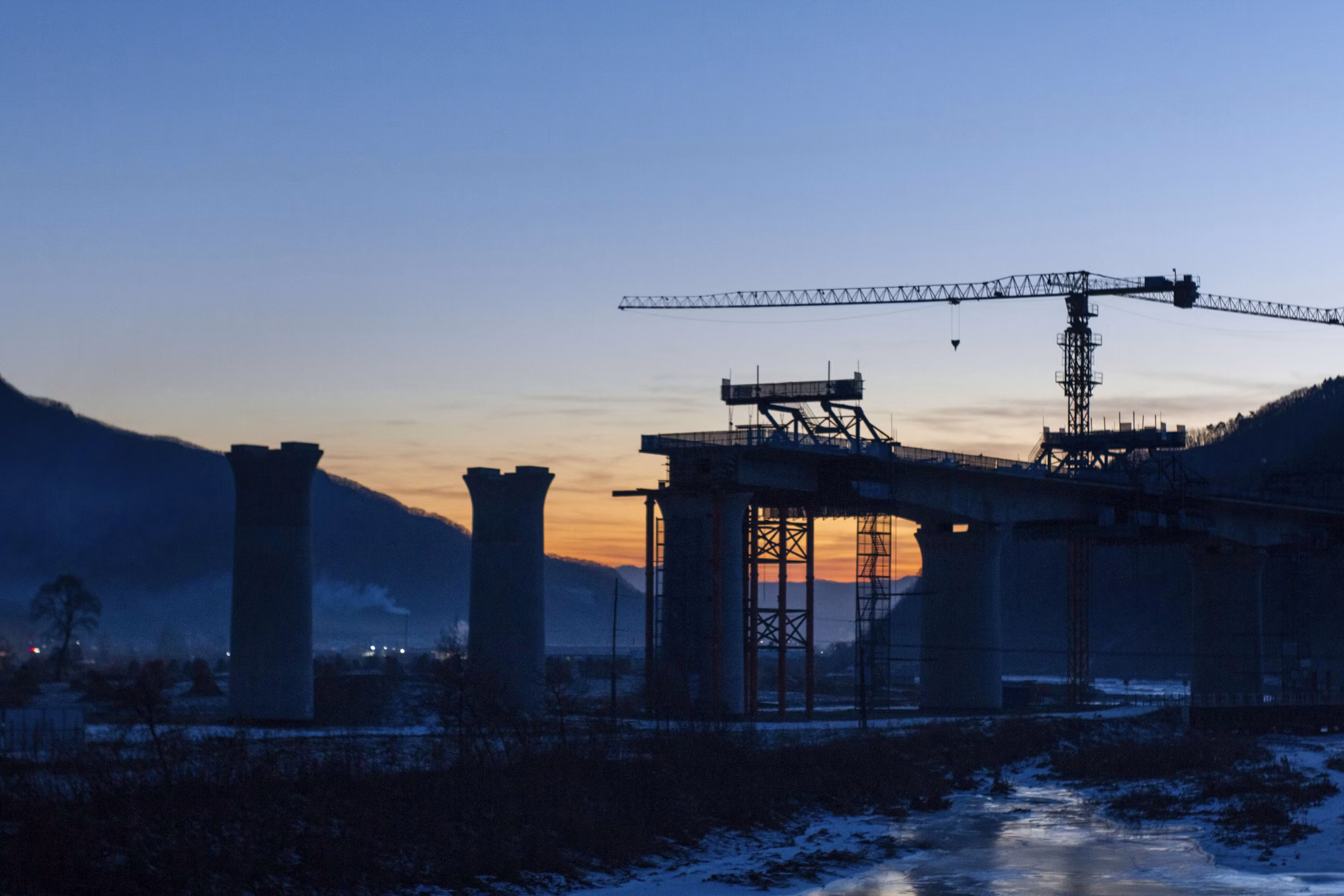 A bridge under construction at dusk, with concrete piers and a tower crane in silhouette.