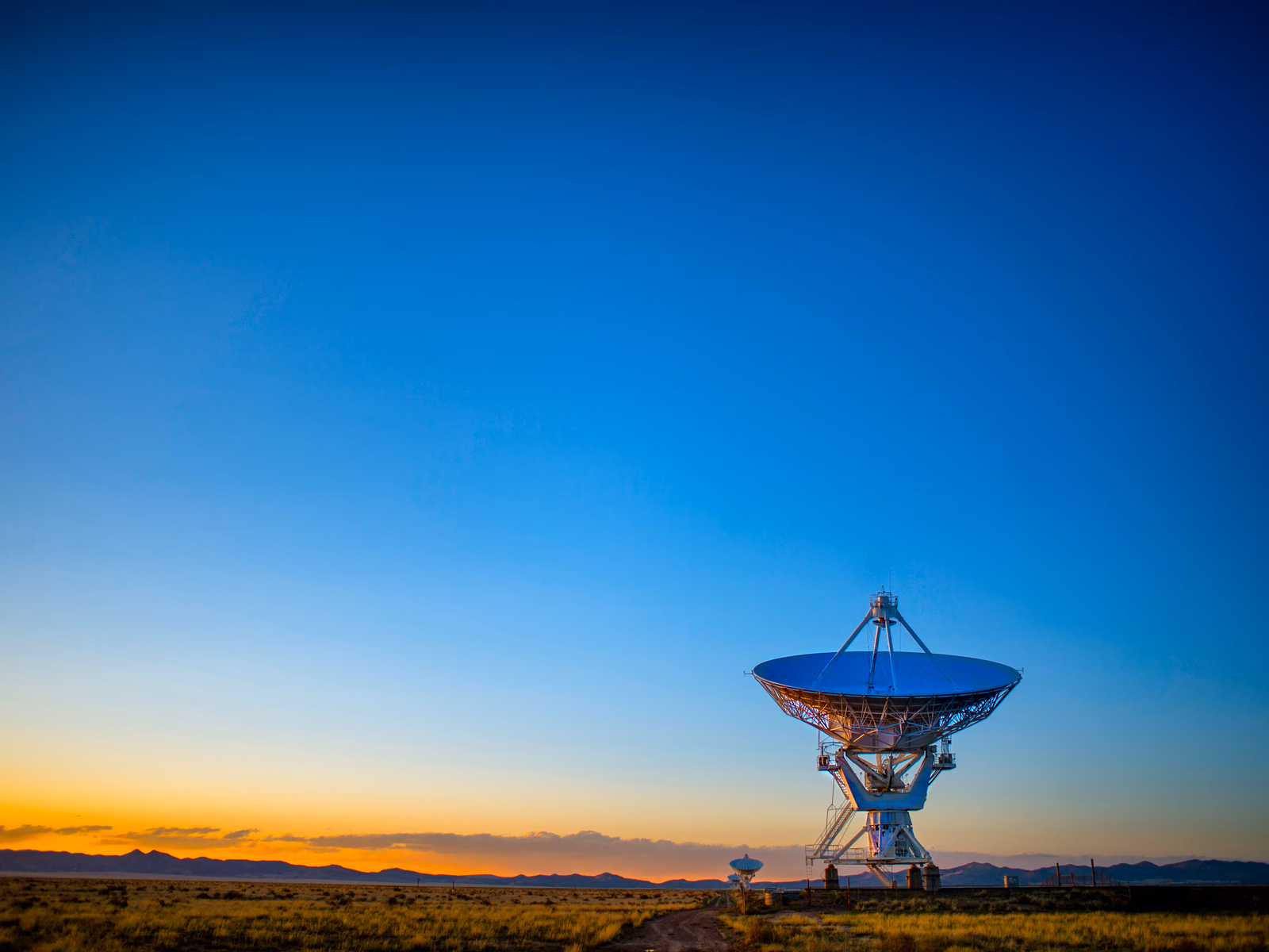 A radio telescope dish on open plains at dusk, its face lit against a deep blue sky above a sunset horizon.